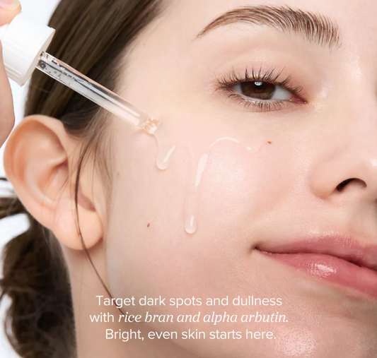 Close-up of a woman's face with droplets of skincare product, text about rice bran and alpha arbutin.