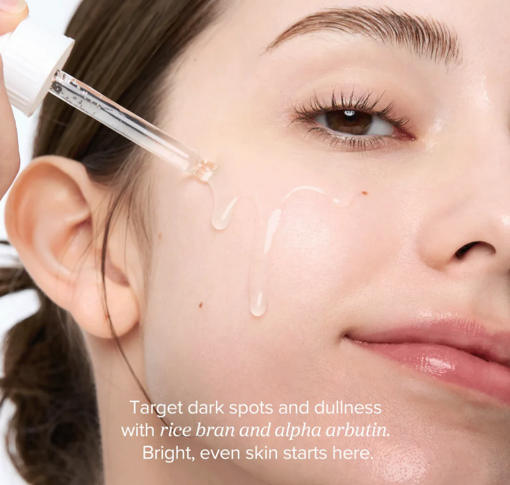Close-up of a woman's face with droplets of skincare product, text about rice bran and alpha arbutin.
